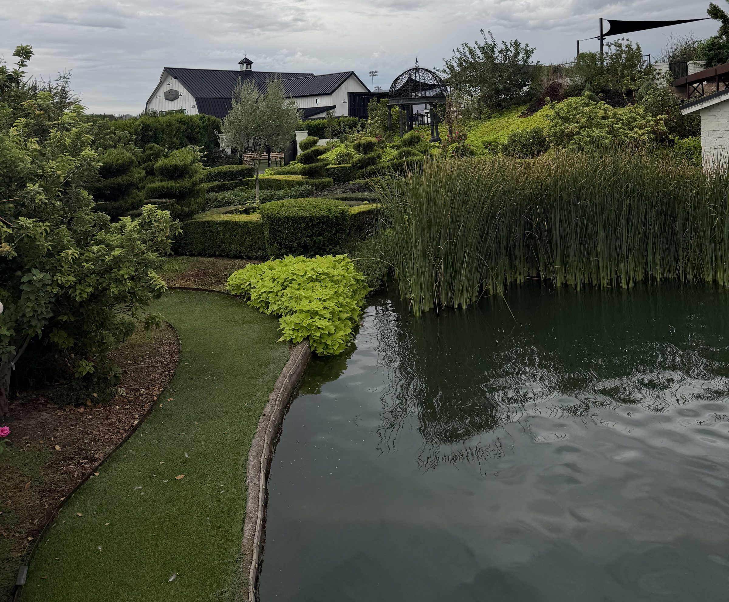 A curved lawn path beside the pond, the millhouse roof and the iron gazebo dome visible in the distance