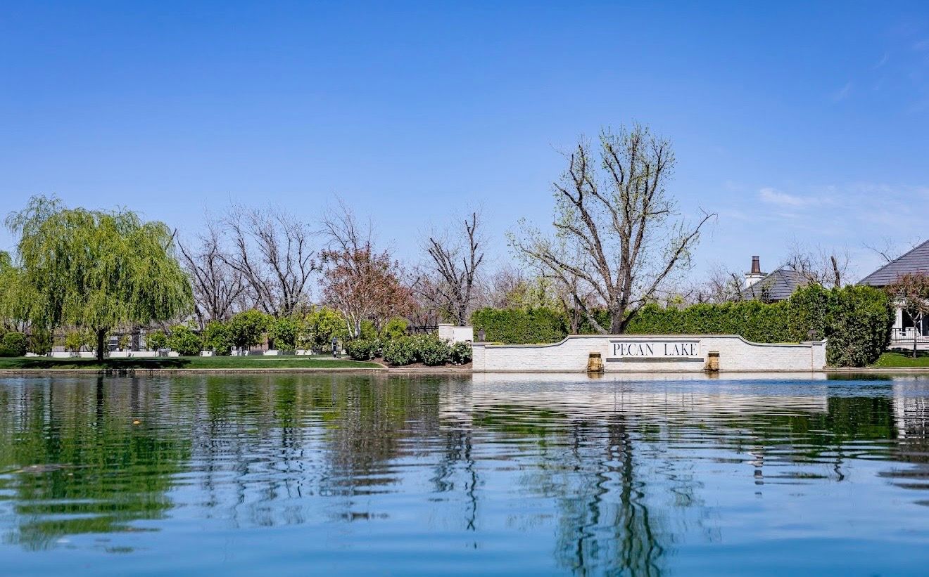 The Pecan Lake sign wall reflected in still water, willows along the bank